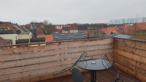 Una terraza de madera con una mesa redonda y sillas de metal, con vistas a una zona residencial con casas de tejados rojos y un edificio lejano.