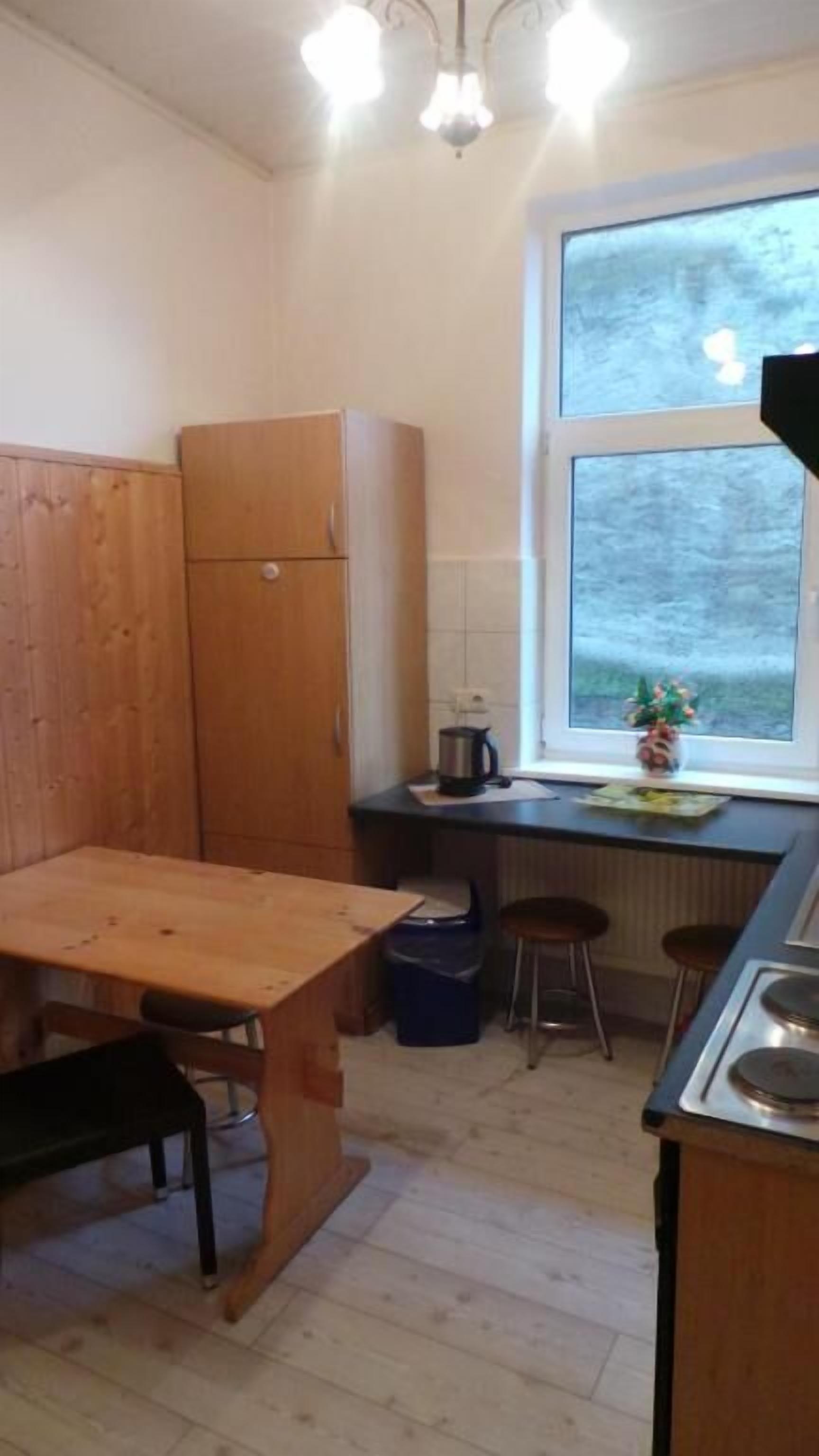 Cocina compacta con una mesa de madera, un taburete pequeño, una tetera y un ventanal con vistas a árboles.