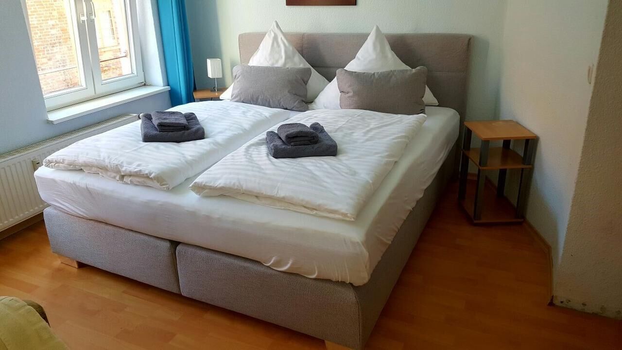 A double bed with white linens and two pillows, a small bedside table, and a window with curtains.