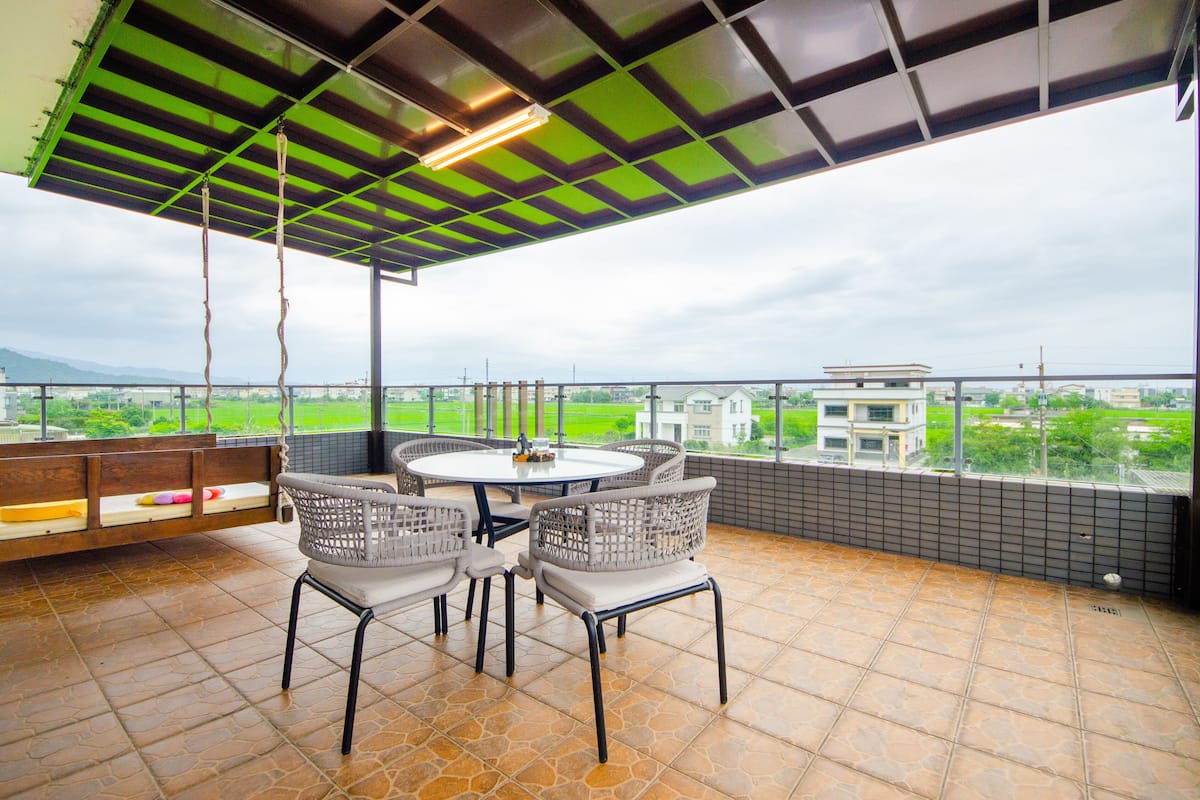 A covered outdoor seating area with a table and chairs, a bench, and a view of buildings and greenery.