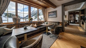 A cozy interior with a large window offering a mountain view, a seating area with a table, and a kitchen visible in the background. - Hotel Montanara (Ischgl)