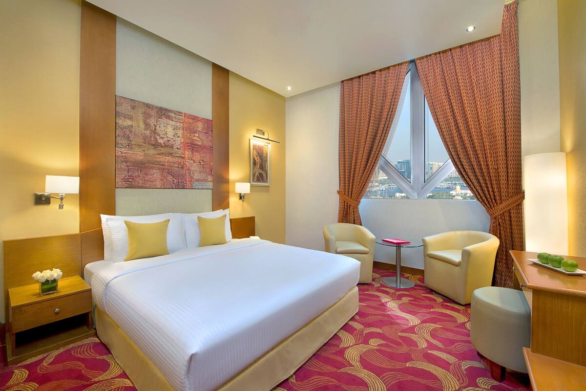 A hotel room with a large bed, two chairs, a small table, and a view of the city through a window.