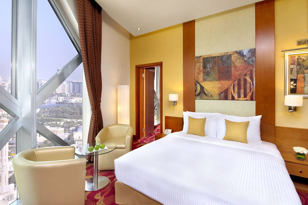 A hotel room with a large bed, a desk with a chair, a view of the city through a large window, and a painting on the wall.