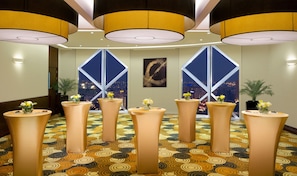 A hotel lobby with a modern design, featuring a patterned carpet, tall cylindrical tables with flower arrangements, and large windows offering a city view.
