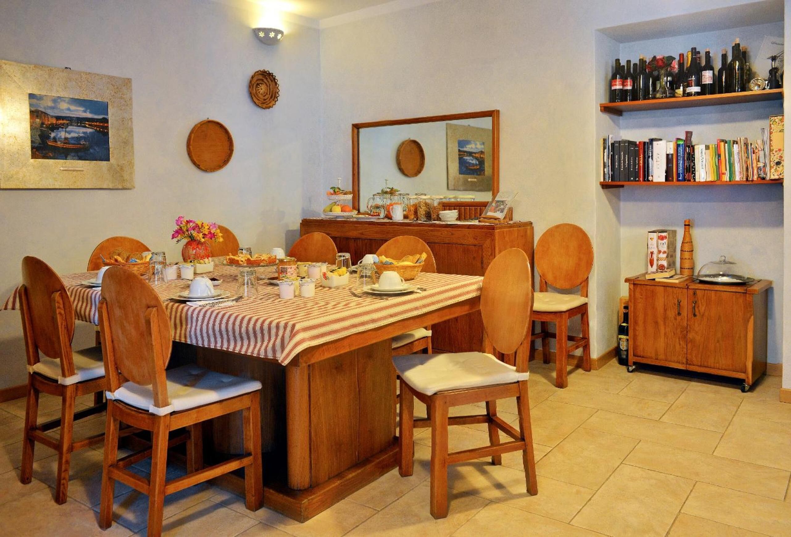 Un comedor con una mesa de madera dispuesta para seis personas, rodeada por seis sillas de madera. Una mesa de buffet en el centro presenta diversos platos y bebidas. A la derecha, una estantería con libros y botellas de vino.