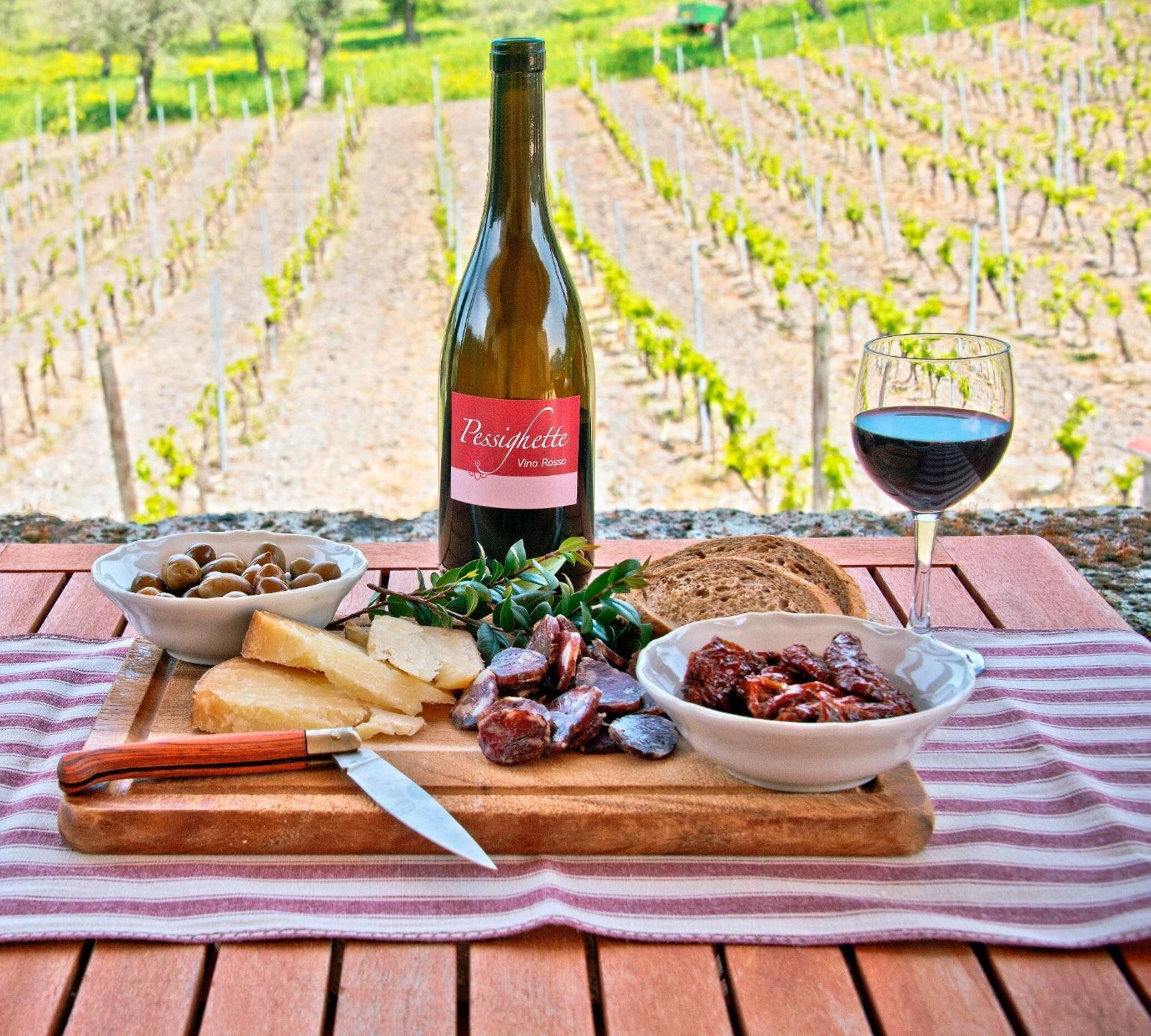 A picnic setup with a bottle of wine, cheese, olives, bread, and charcuterie on a wooden table with a vineyard in the background.