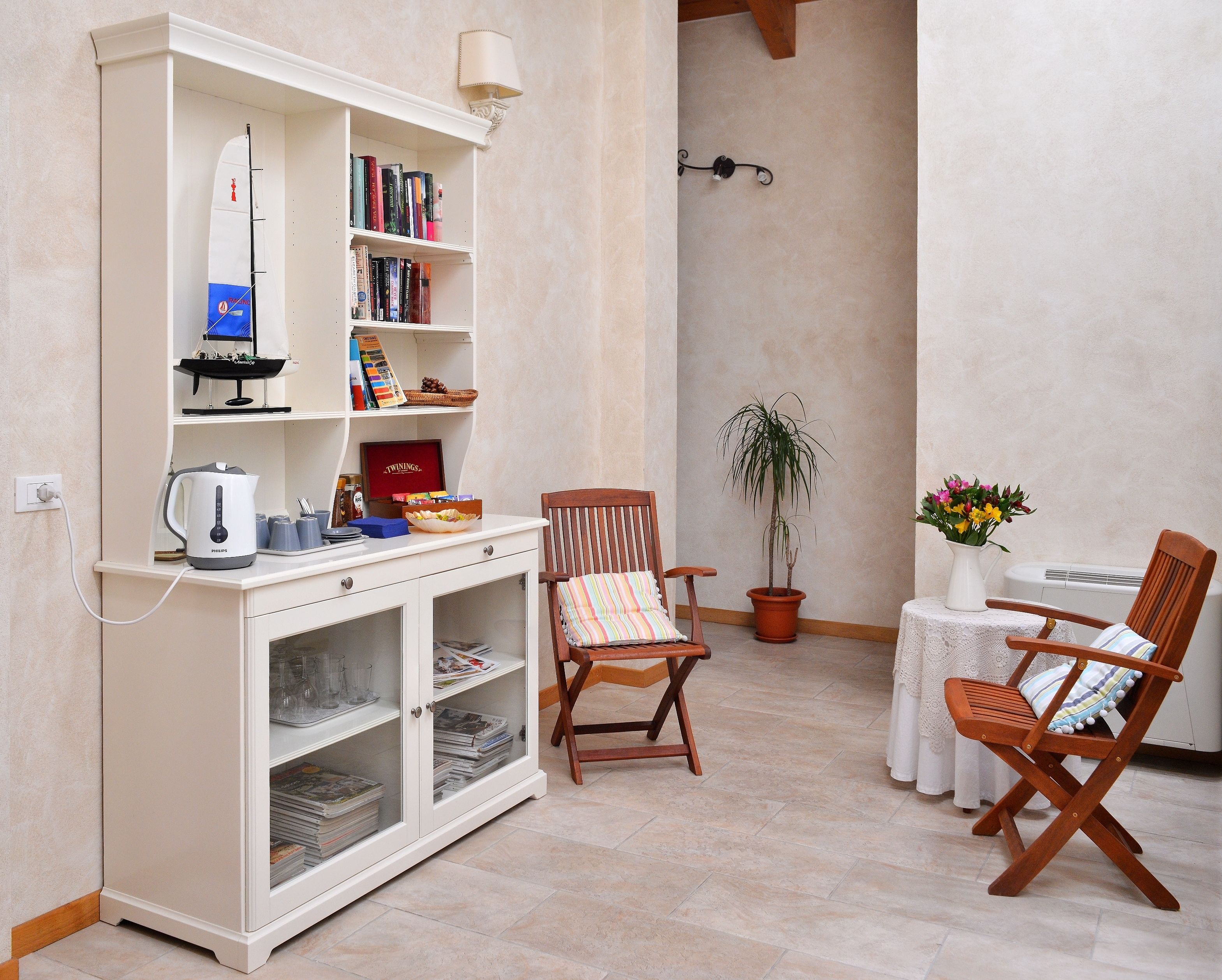 A cozy living area with a white cabinet, bookshelves, a coffee table, two wooden chairs, a potted plant, and a small table with a vase of flowers.