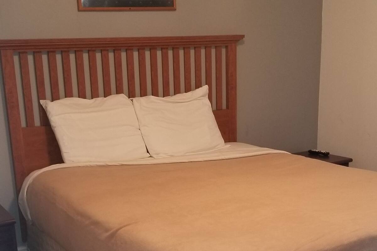 Uma cama bem arrumada com cabeceira de madeira, um quadro na parede e um chão coberto por carpete escuro.