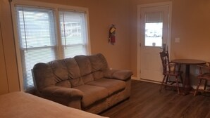 A living room with a brown sofa, a wooden dining table with chairs, a white door, and a window with blinds. - Rip Van Winkle Motel (Plattsburgh)