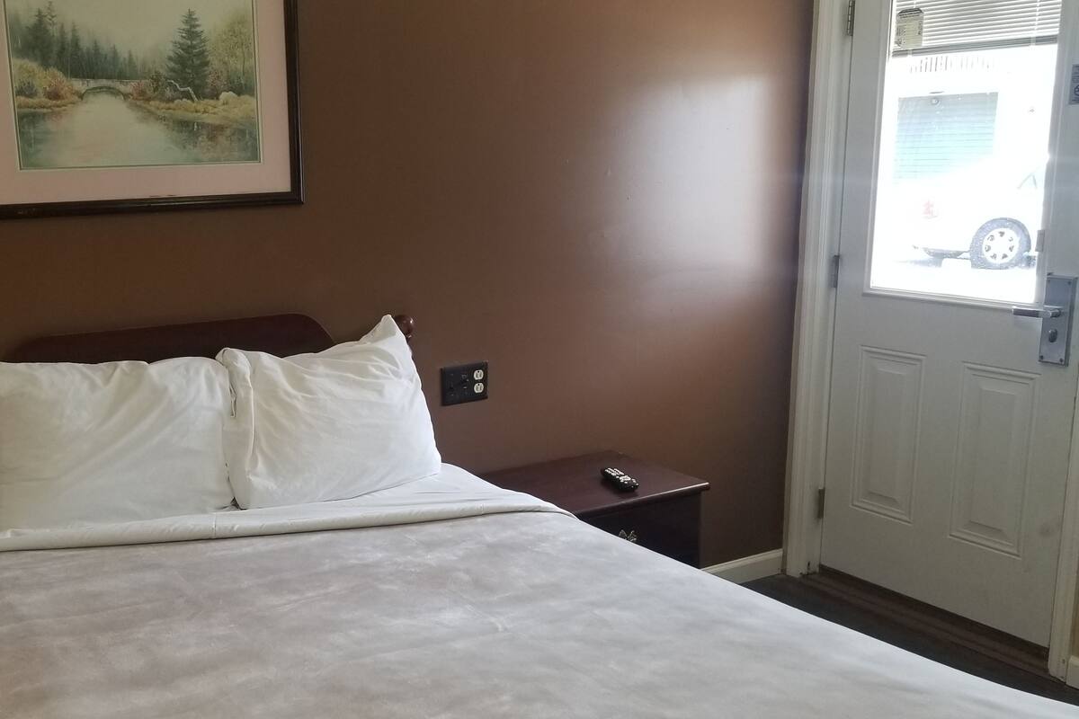 Uma cama bem arrumada com lençóis brancos, uma cabeceira de madeira, uma pequena mesa de cabeceira com um controle remoto, e um quadro de uma ponte sobre um rio na parede.