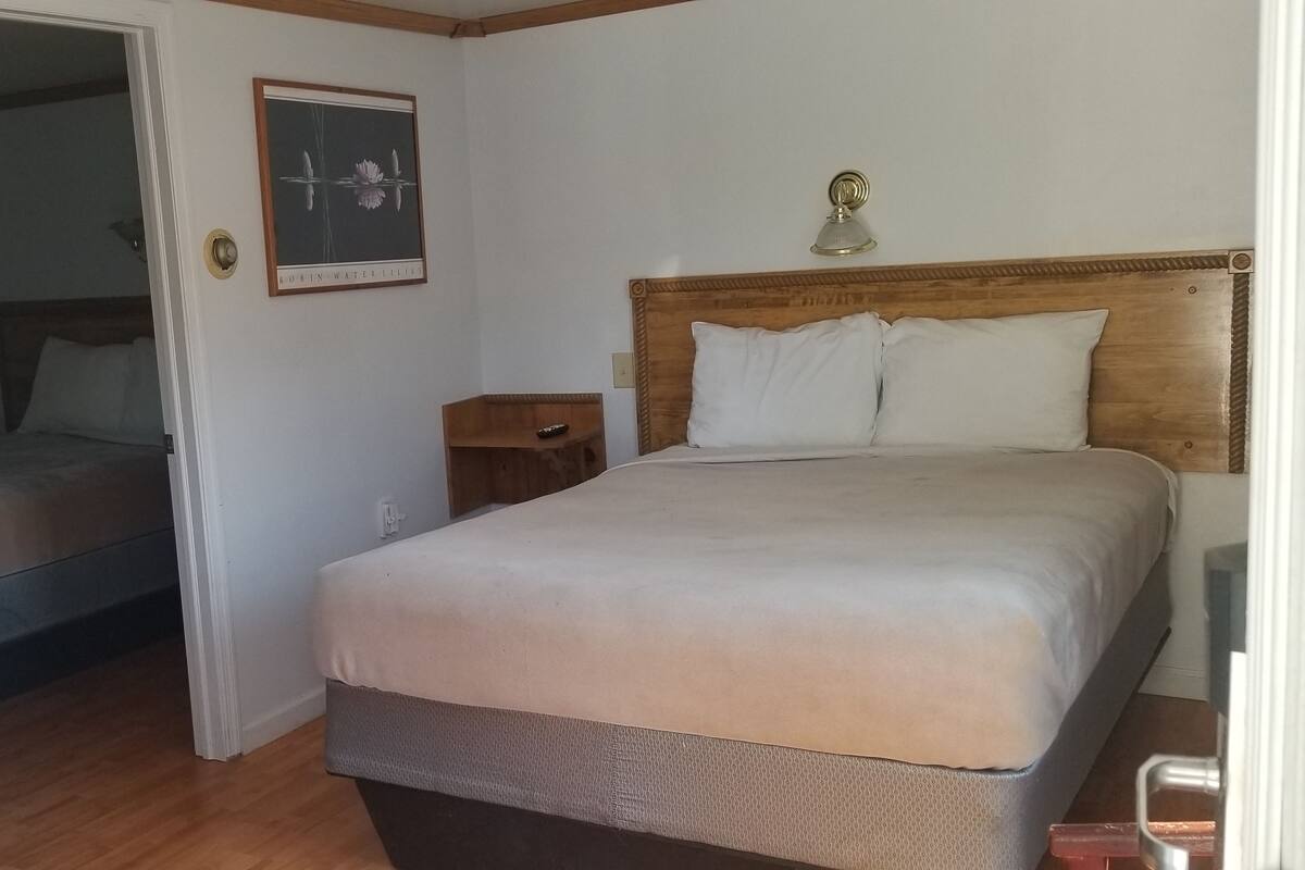 Quarto com cama, mesa de cabeceira, quadro na parede e porta para outro cômodo.