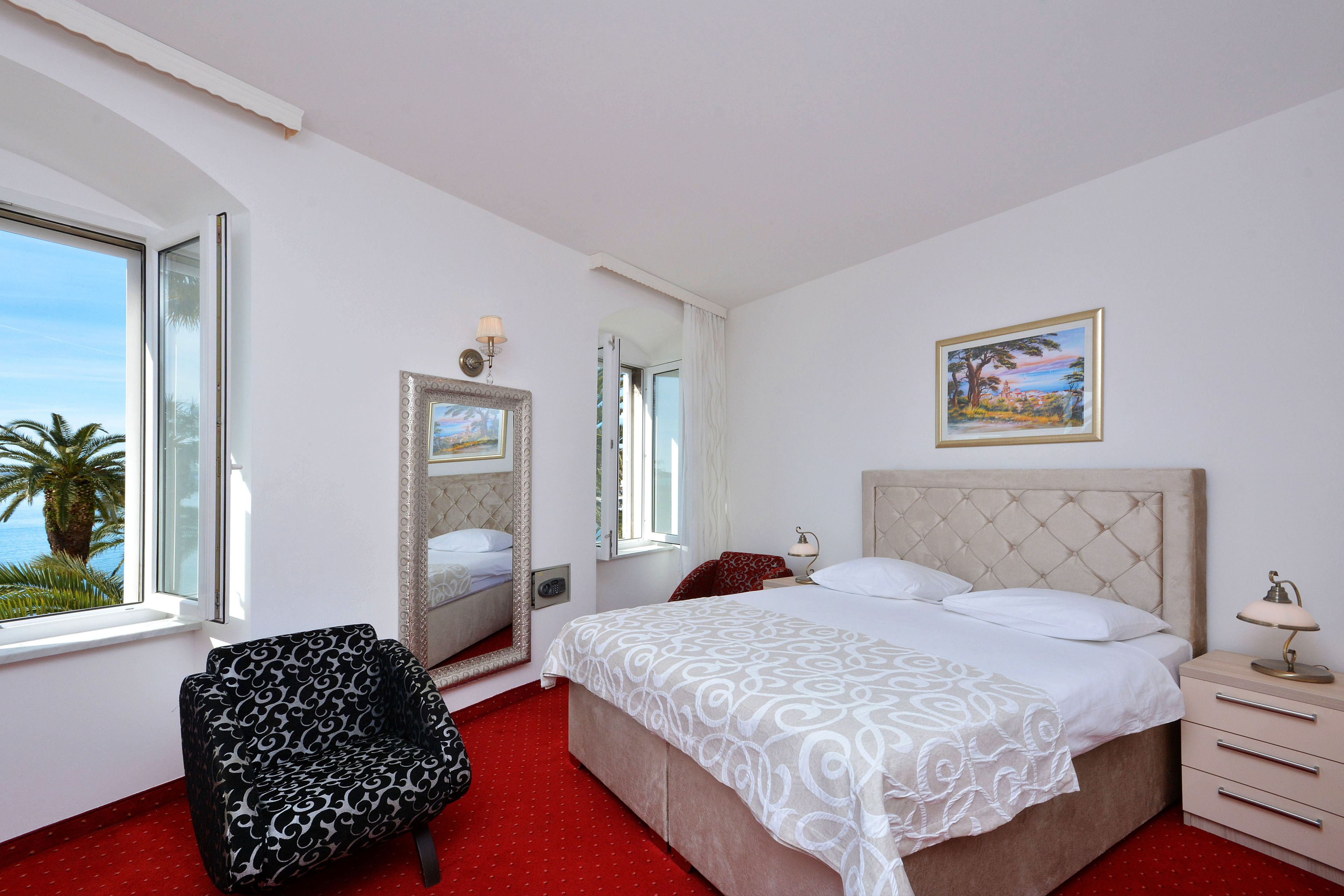 A bedroom with a bed, a chair, a nightstand, a lamp, a painting, and a window with a view of palm trees.