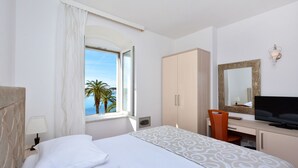 Una habitación de hotel con cama, escritorio, televisión, armario y ventana con vista a palmeras y al mar.