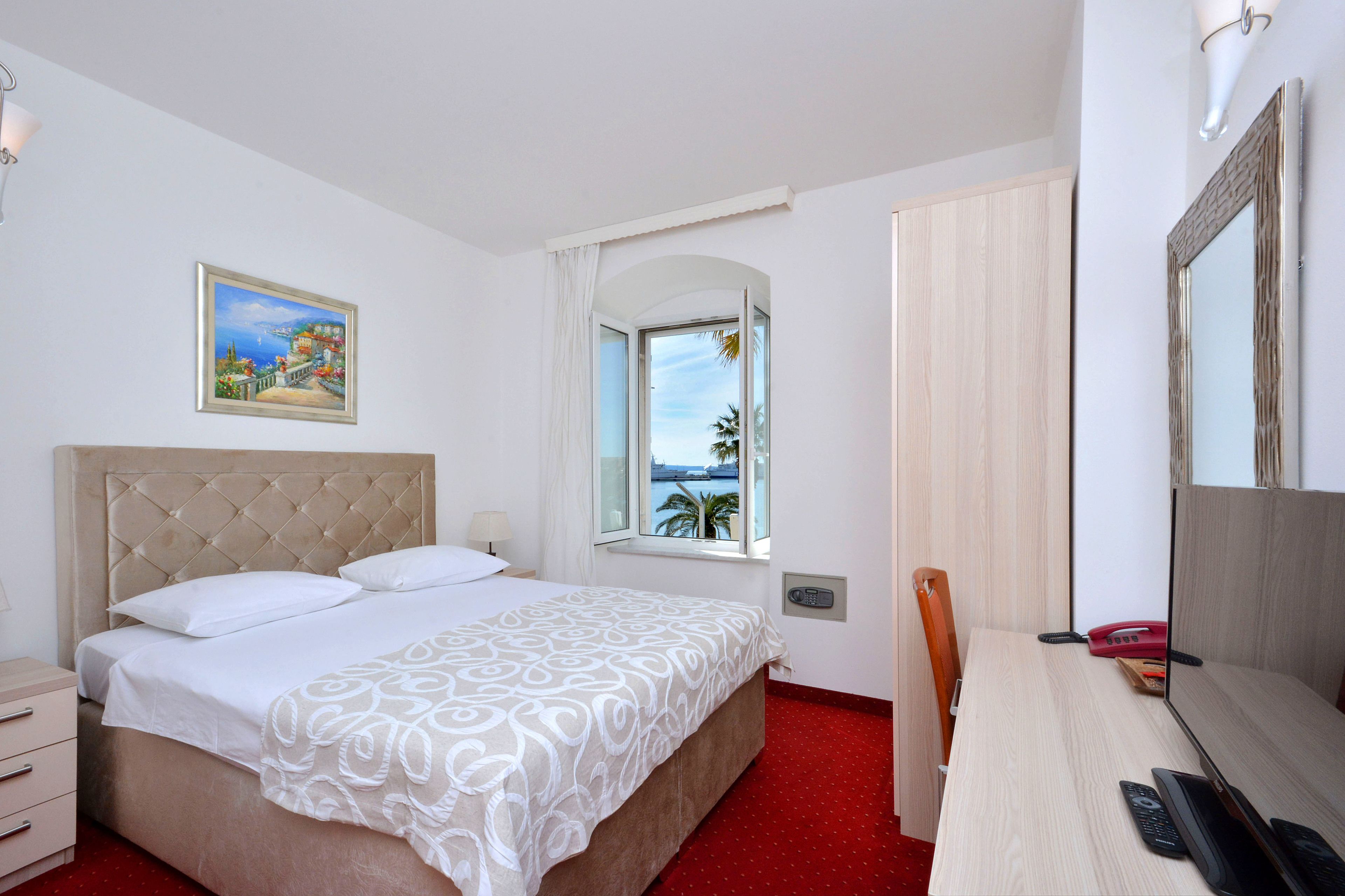 A hotel room with a bed, a desk, a chair, a window with a view, a painting on the wall, and a telephone.