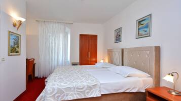 A hotel room with a large bed, a wooden wardrobe, a desk with a chair, and a window with curtains.