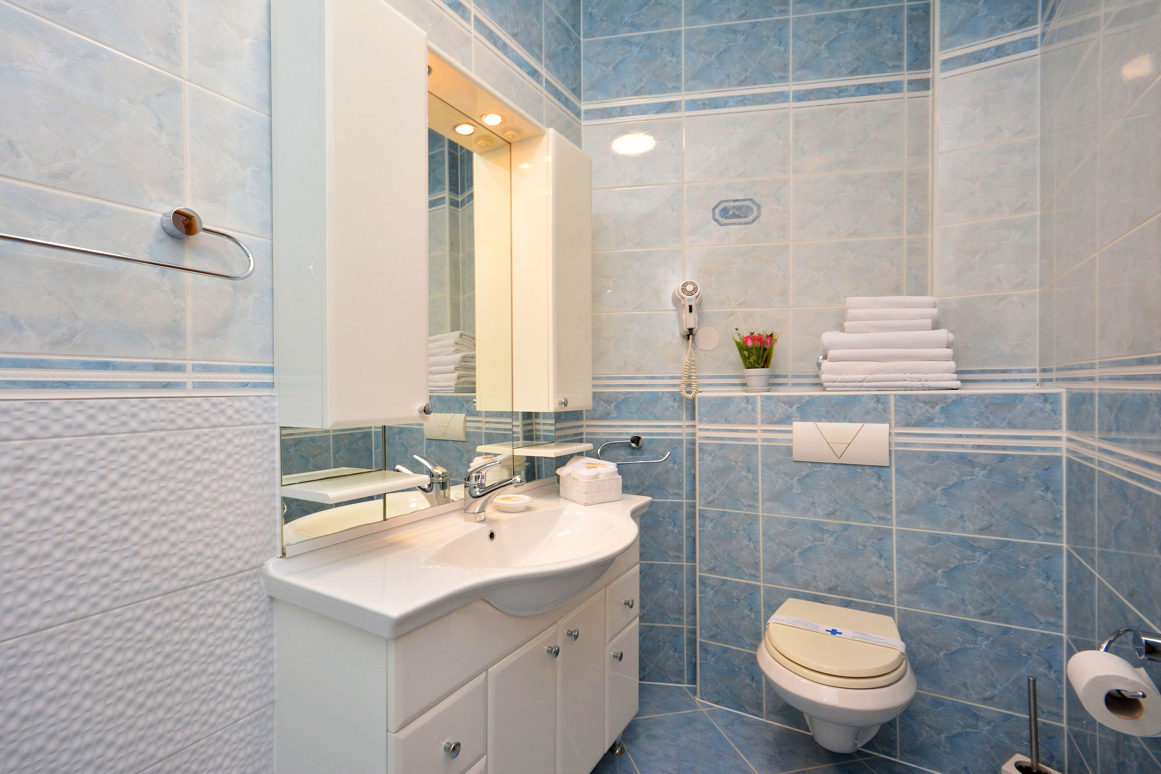 Un baño con lavamanos blanco, espejo y retrete. Hay un perchero con toallas dobladas y una pequeña planta en maceta sobre el mostrador.