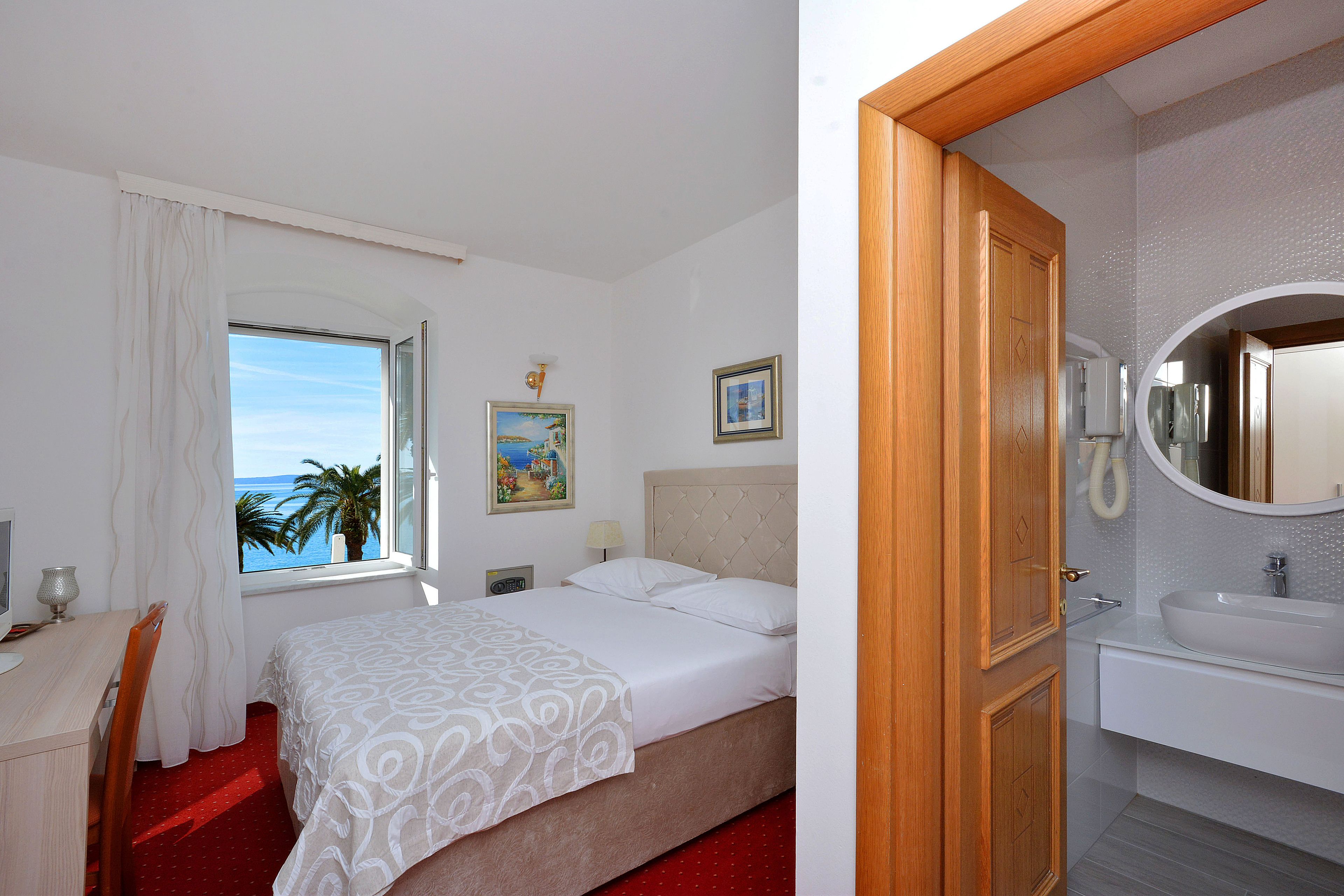 A hotel room with a bed, a desk, a window with a view of the sea, a bathroom with a sink and mirror.