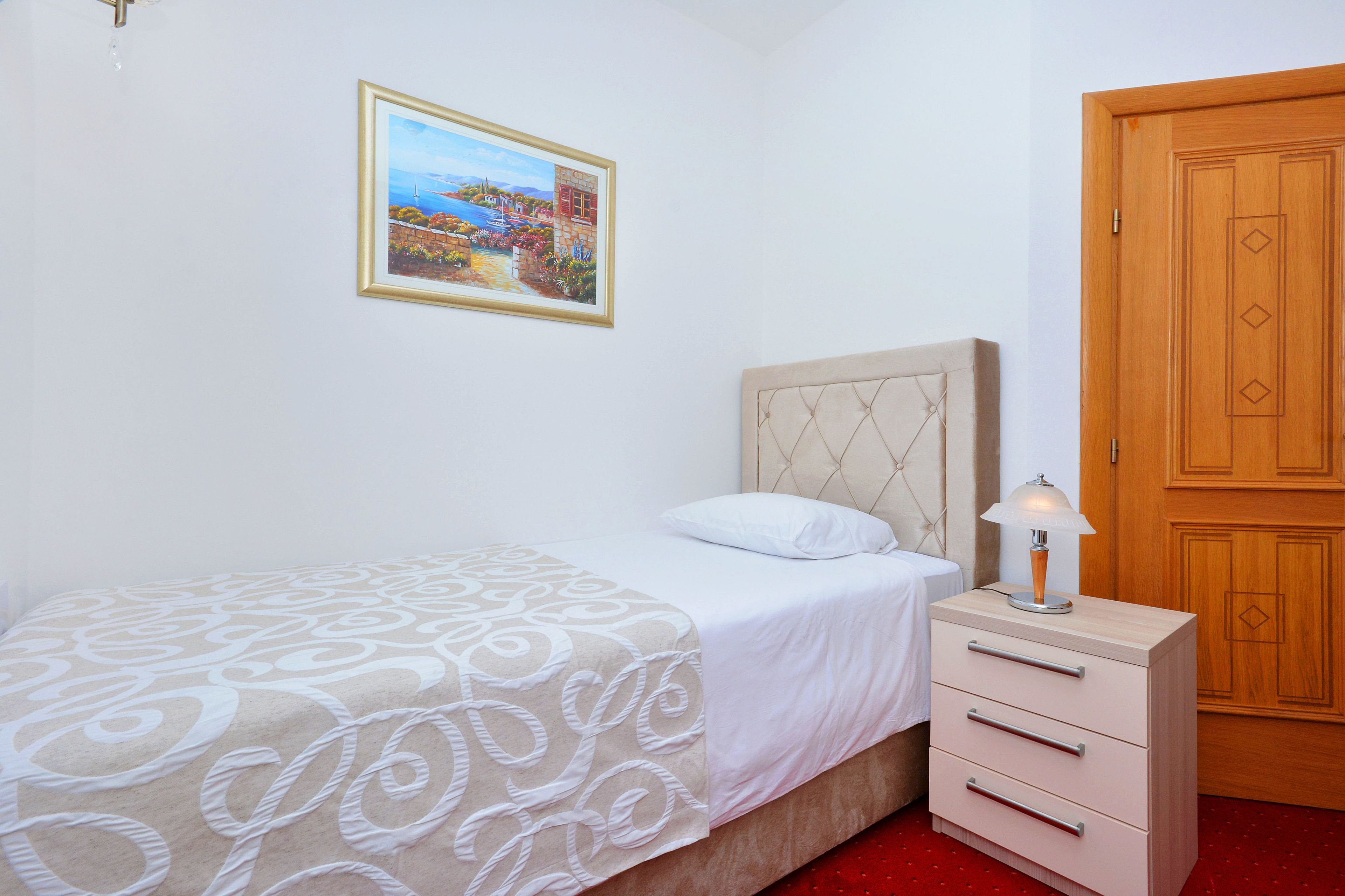 A bedroom with a bed, a bedside table with a lamp, a wooden wardrobe, and a framed painting on the wall.