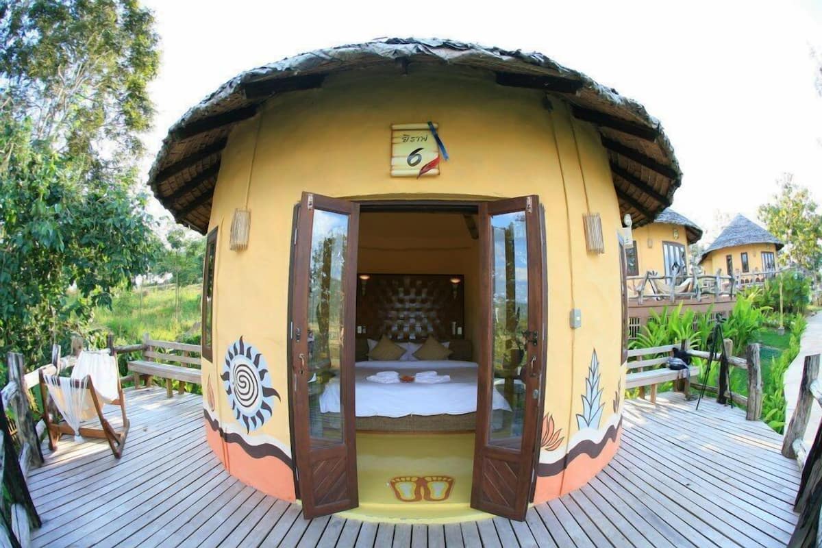A circular, single-room structure with a thatched roof, a bed, and a small balcony.
