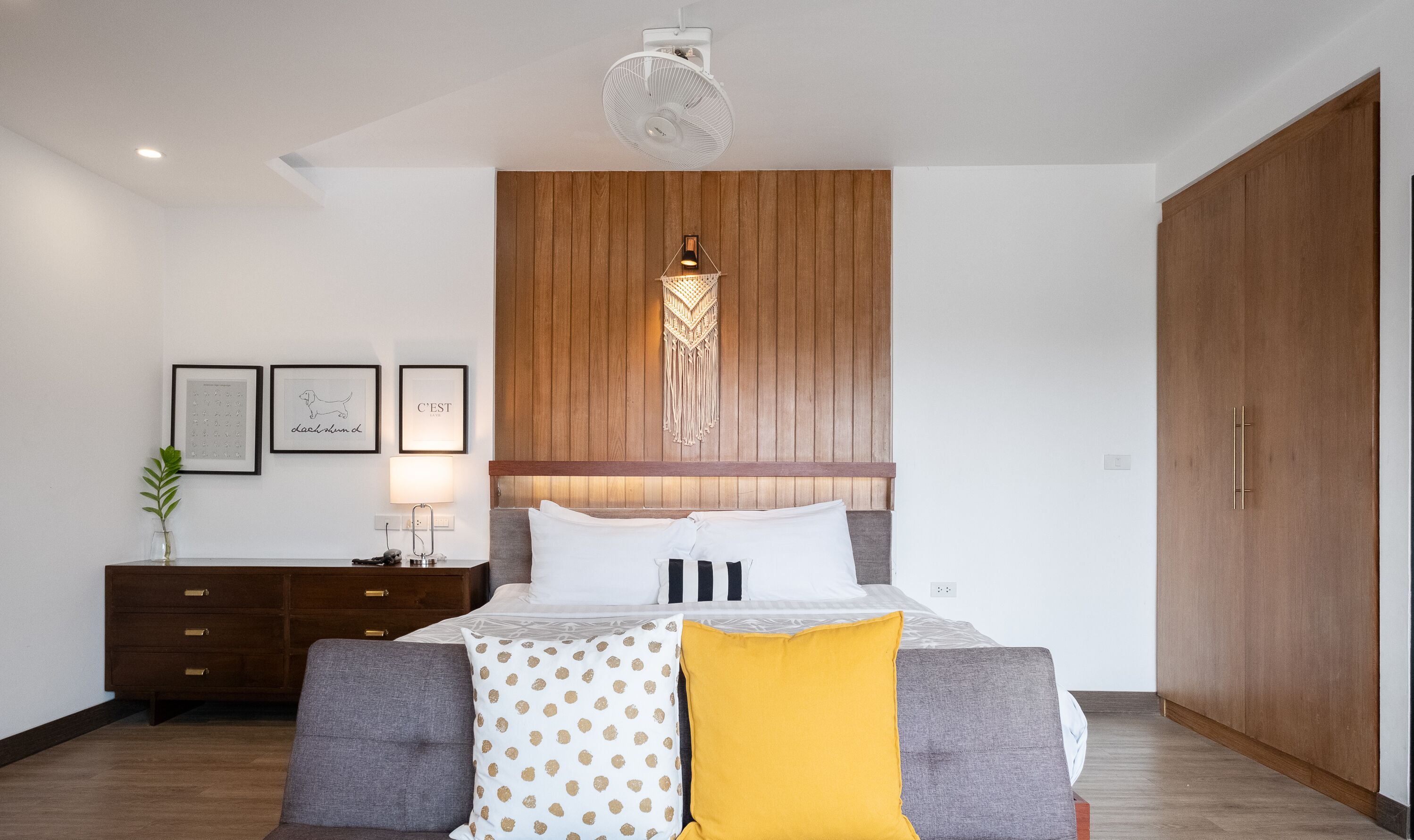 Et moderne soveværelse med sengegavl i træ, seng med hvide og mønstrede puder, et natbord med lampe og en stor garderobe.