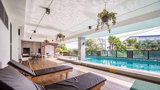 A poolside area with lounge chairs, a brick wall, and a view of a building and trees. - The Corner Lodge (Pattaya)