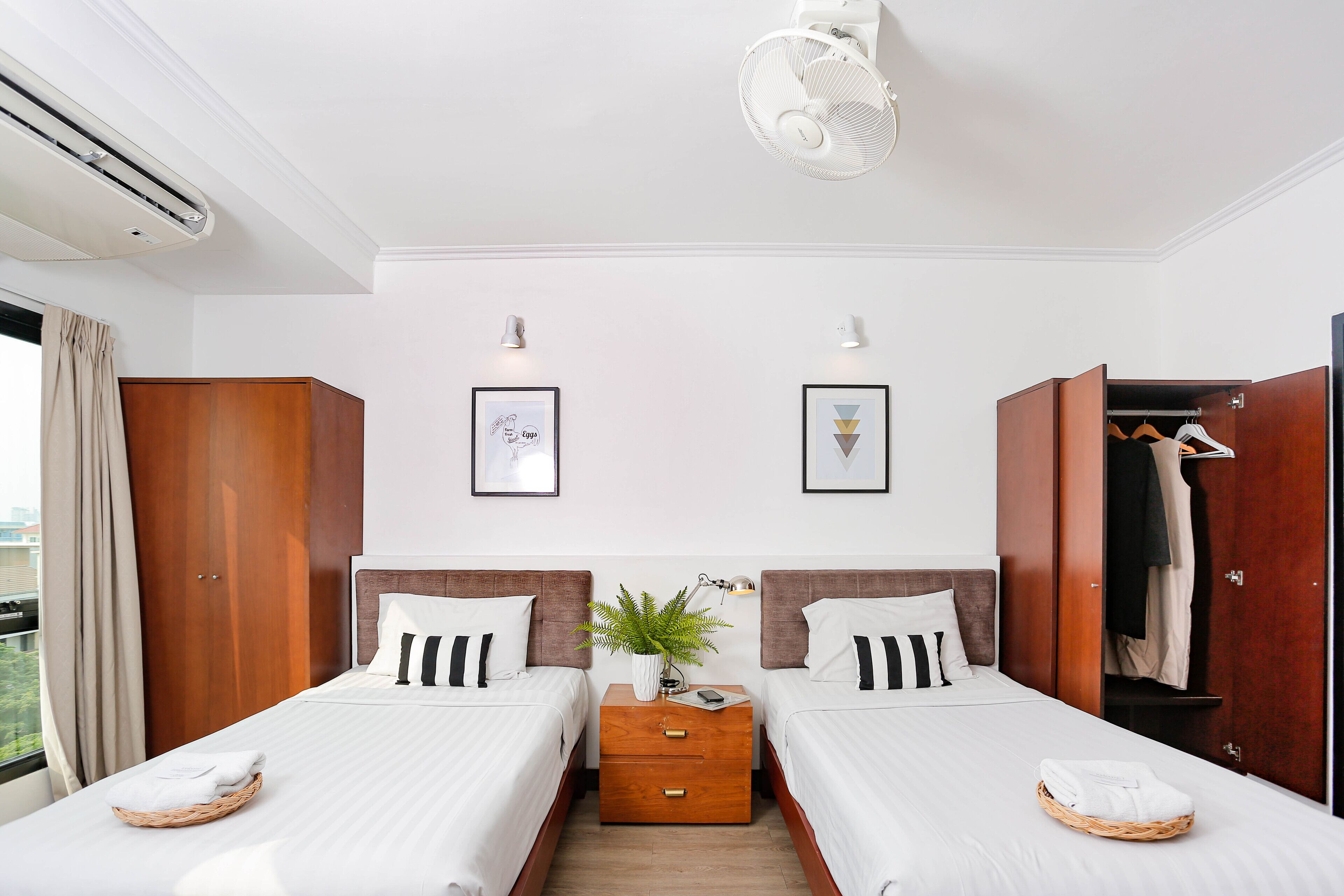 Habitación de hotel con dos camas, un armario de madera, una mesita con una planta y una ventana con cortinas.