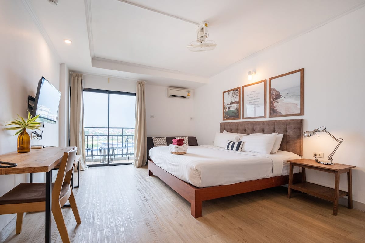 A hotel room with a bed, bedside table, desk, chair, and a potted plant.