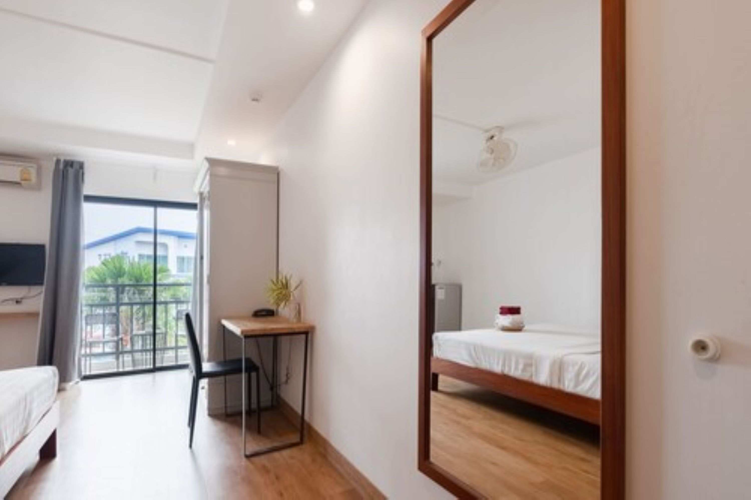 A hotel room with a bed, a desk, a chair, a mirror, and a window with a view of buildings and greenery.