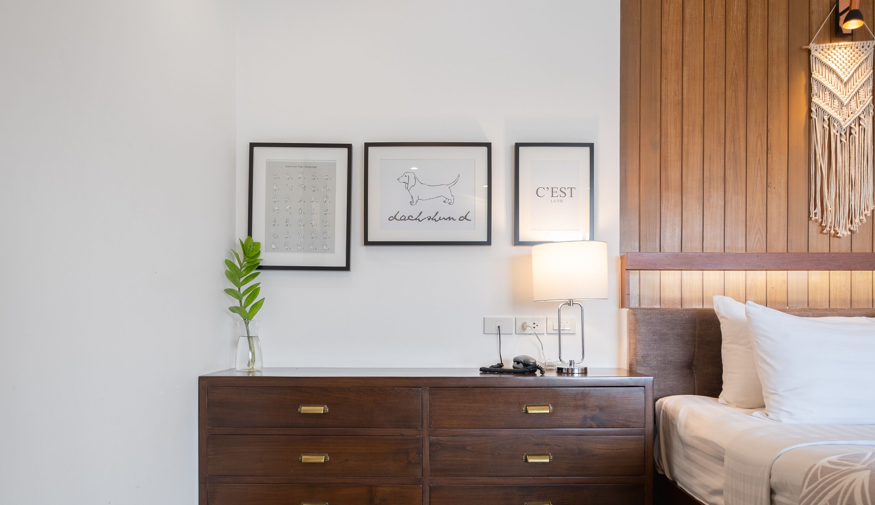 A neatly arranged hotel room with a wooden headboard, a bedside table with a lamp, and three framed pictures on the wall.