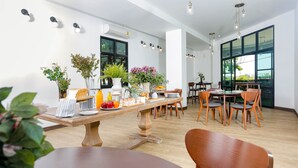 A spacious dining area with wooden tables and chairs, large windows, and various plants. - The Corner Lodge (Pattaya)