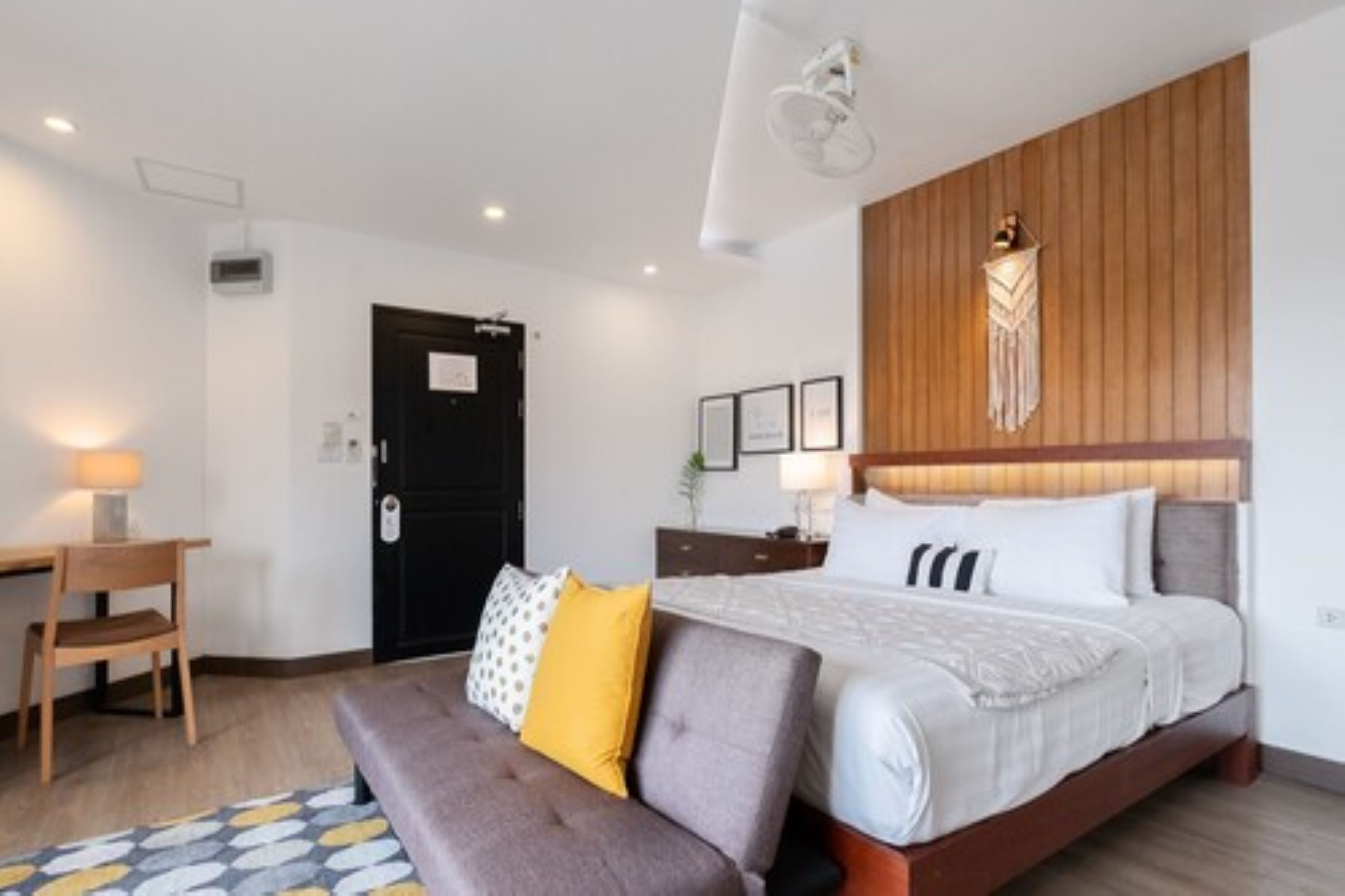 A modern hotel room with a large bed, a wooden headboard, a desk with a chair, and a small table with a lamp.