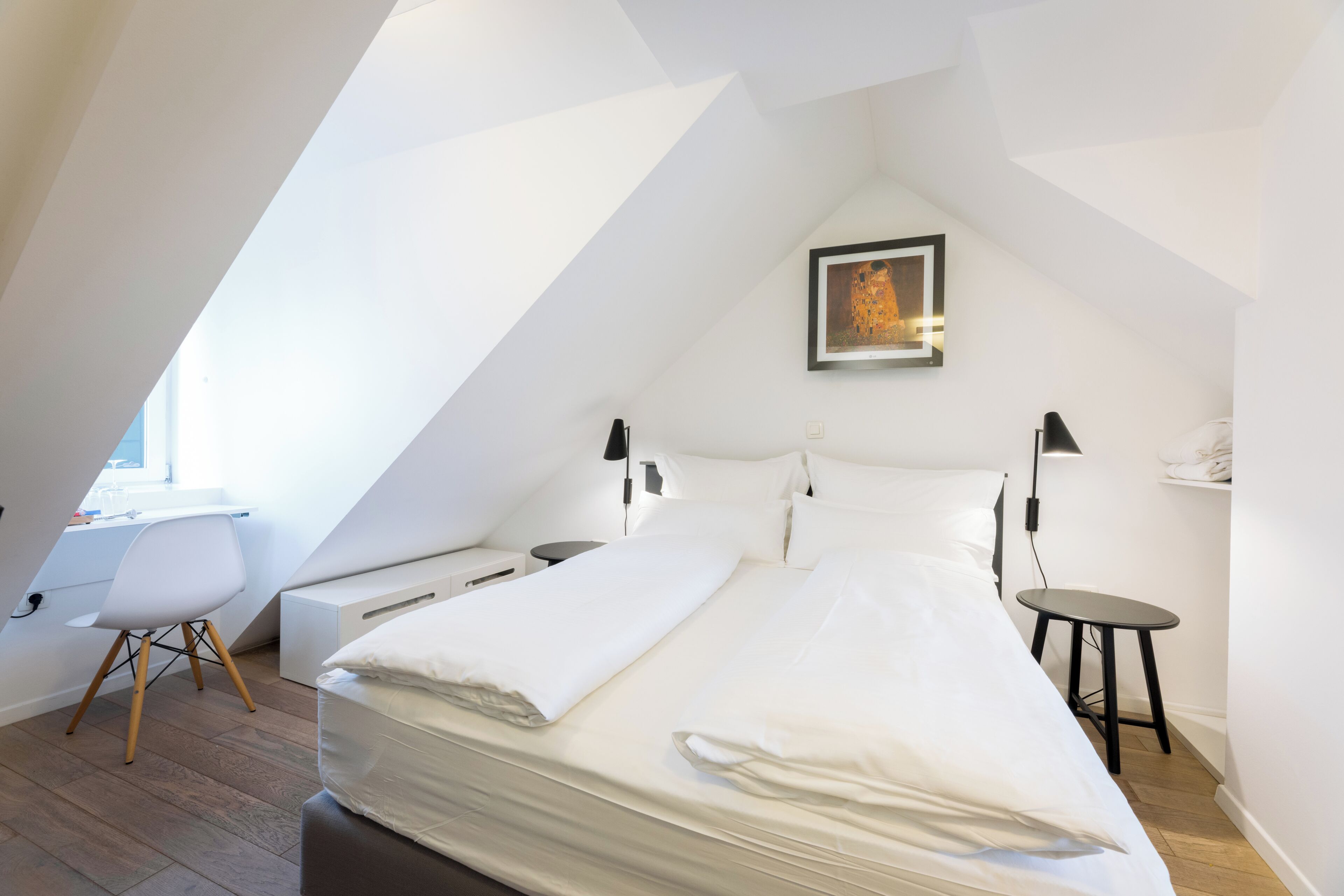 A modern, well-lit room with a bed, bedside tables, a desk, and a chair.