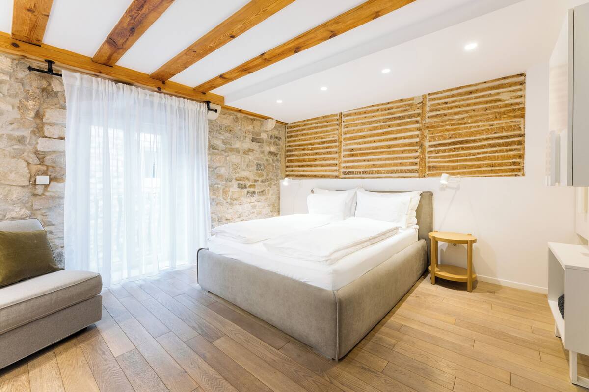 A bedroom with a bed, a wooden headboard, a stone wall, a wooden floor, and a window with curtains.