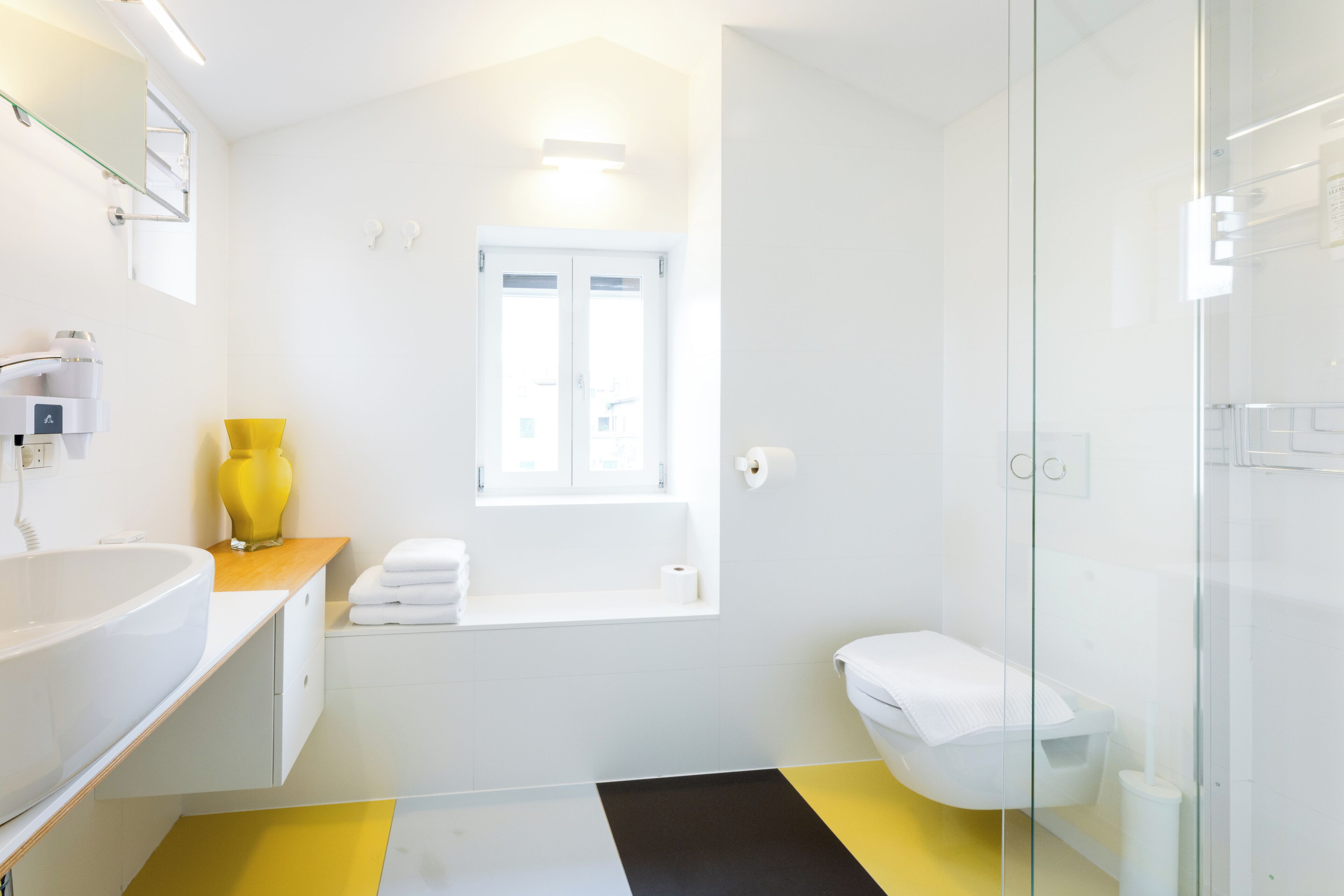 Een moderne badkamer met een witte wastafel, een gele vaas, een wit toilet en een douchecabine van glas.