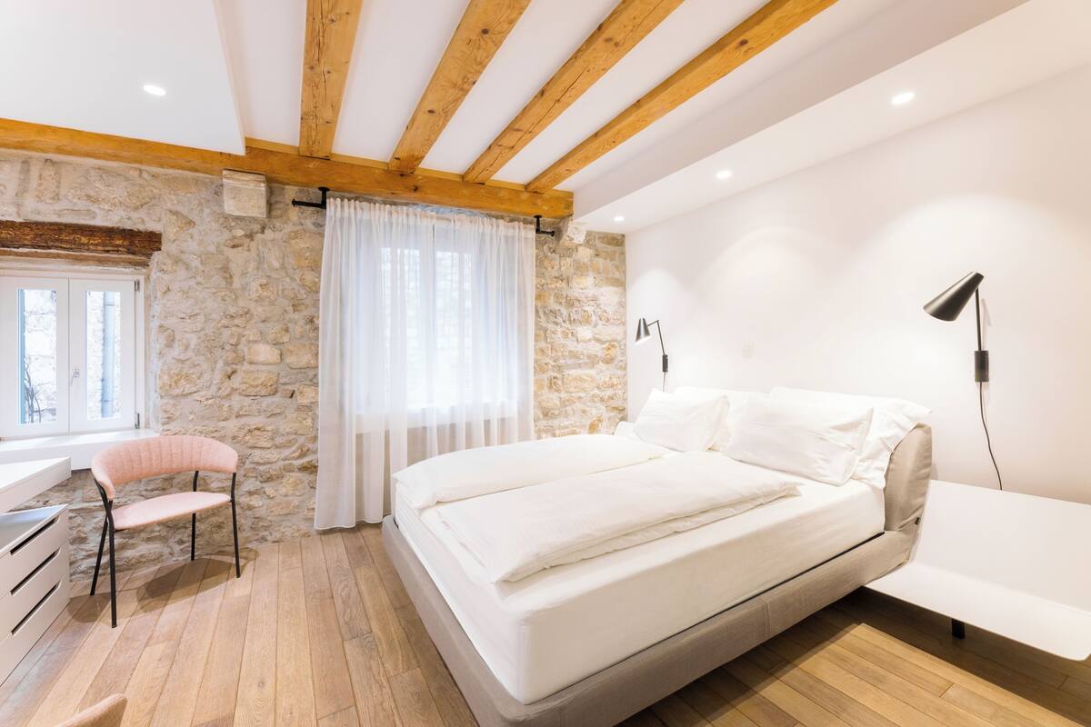 A bedroom with a stone wall, wooden beams, a bed, a chair, and a desk.