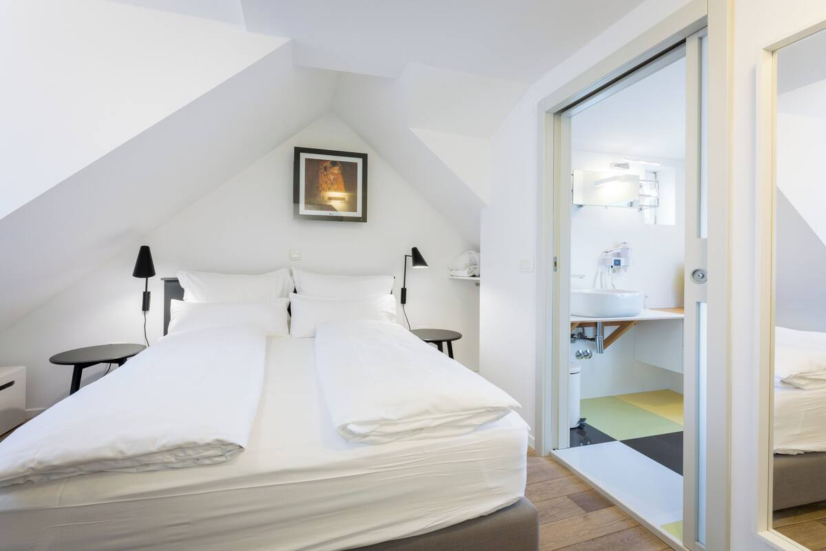A modern hotel room with a bed, bedside tables, a desk, a chair, a mirror, and a bathroom with a sink and toilet.