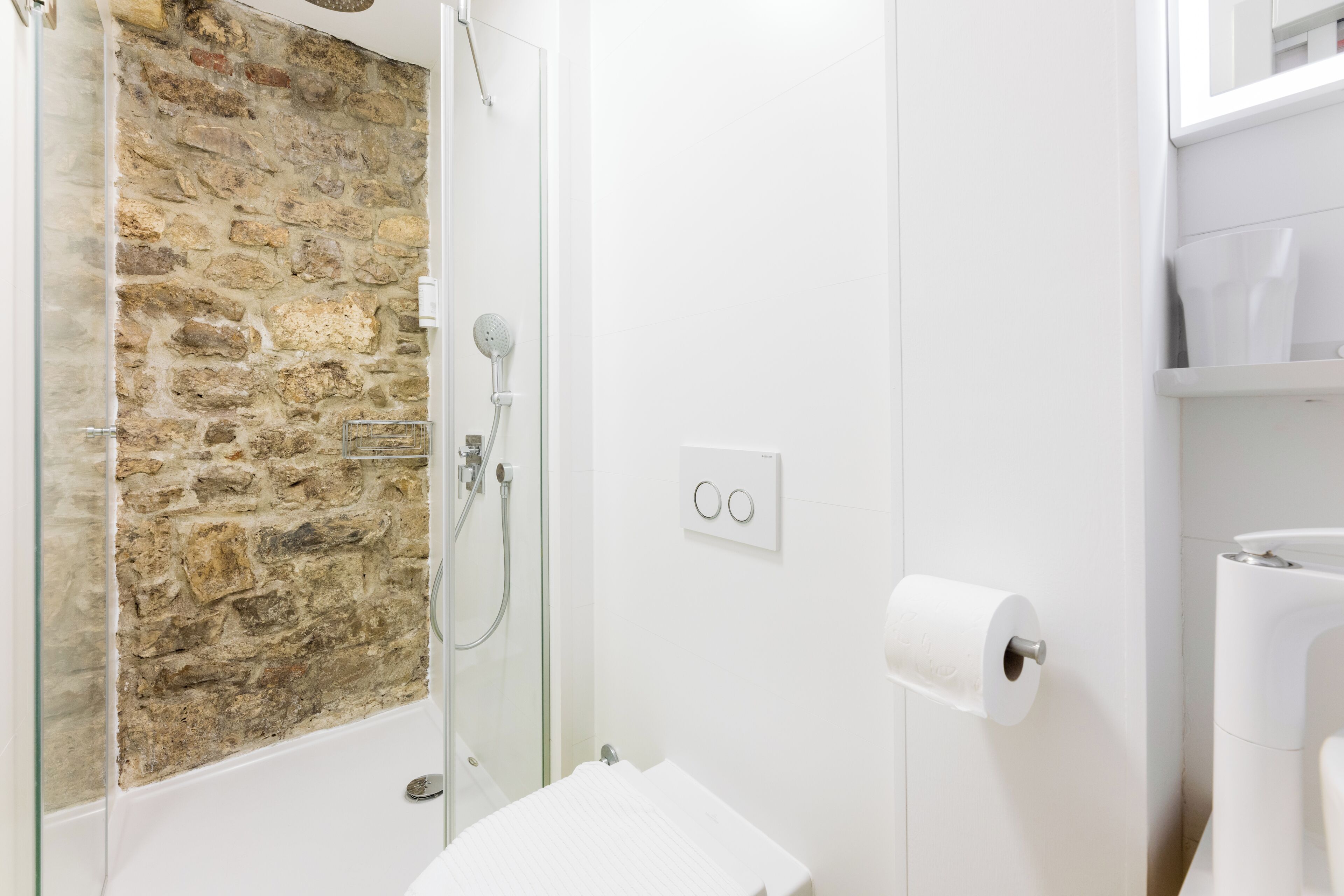 Un baño con pared de piedra, cabina de ducha de vidrio, inodoro y lavamanos blanco.