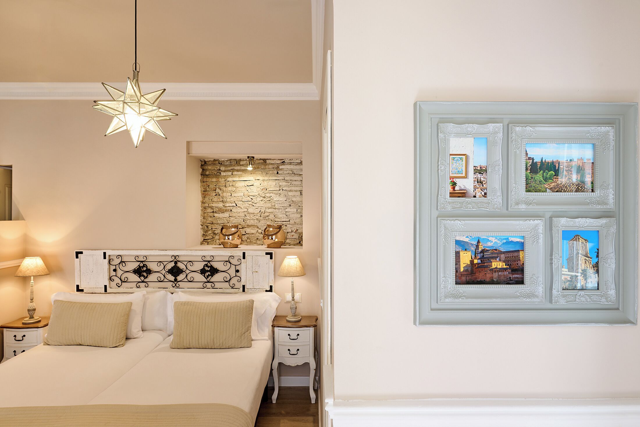 A bedroom with a star-shaped light fixture, a stone wall, a bed with white bedding, two bedside tables with lamps, and framed pictures on the wall.