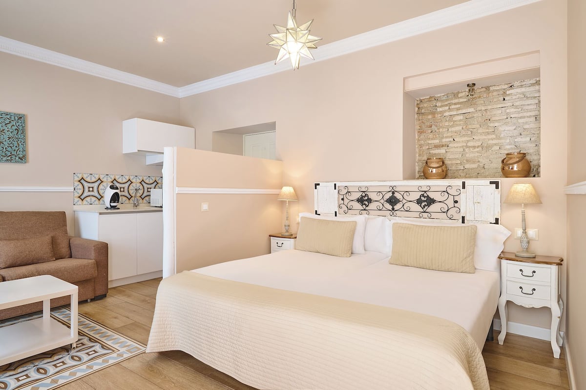 A bedroom with a large bed, bedside tables, a nightstand, a dresser, a hanging light fixture, and a kitchen area in the background.