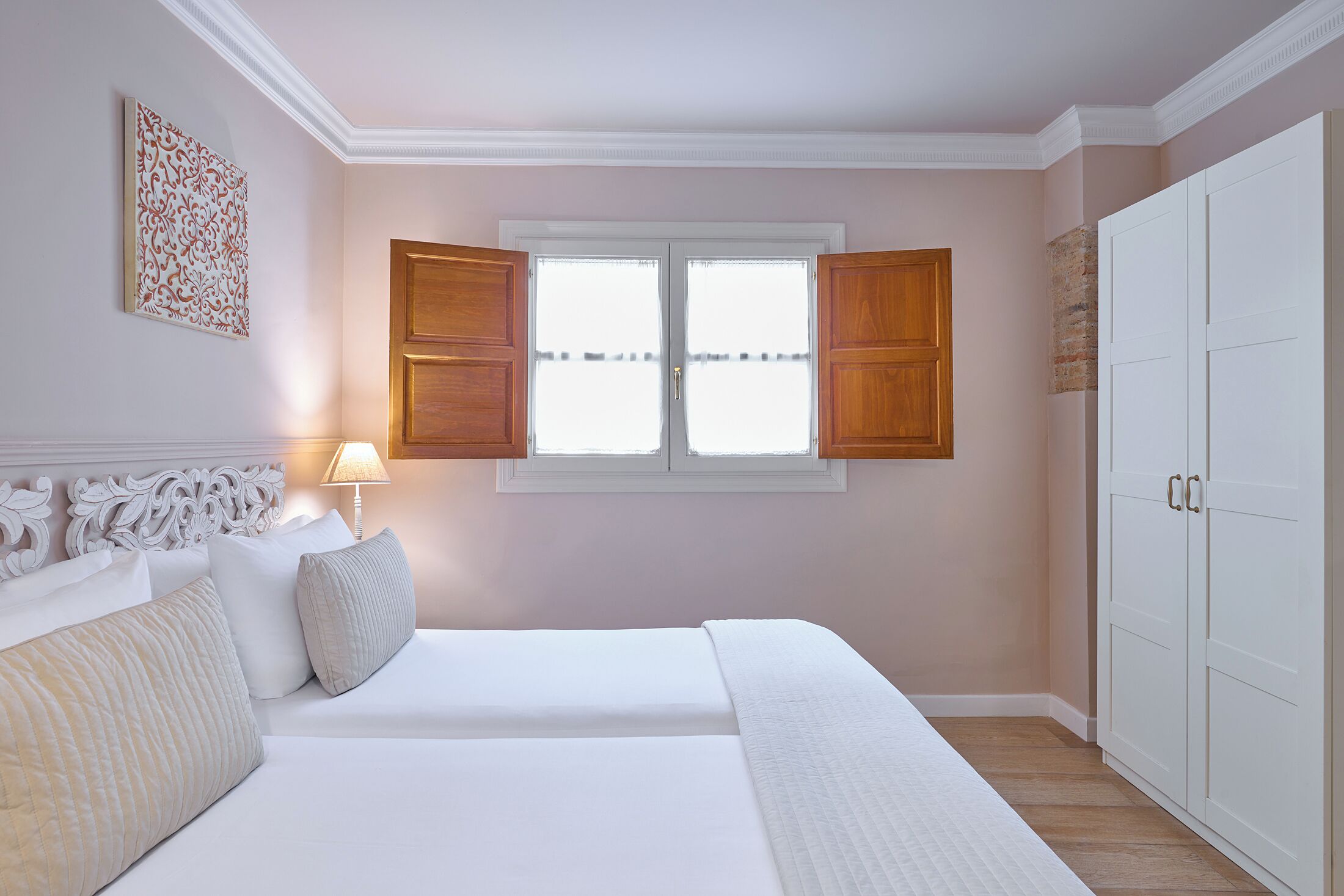 A bedroom with a bed, a nightstand, a lamp, a window with shutters, and a wardrobe.