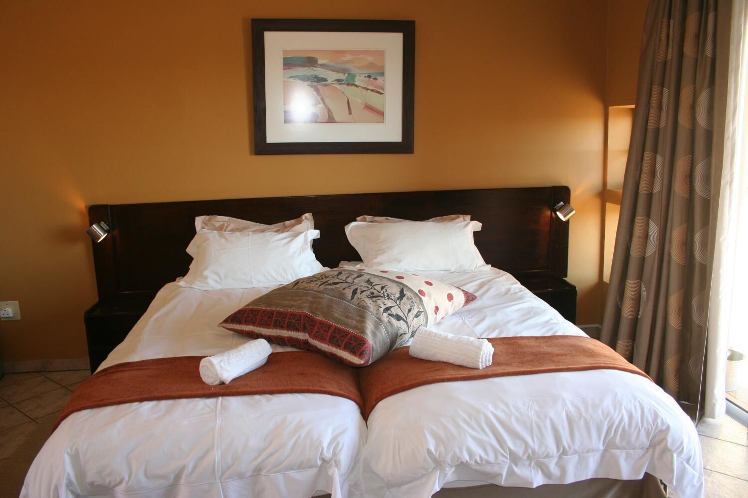 A double bed with white linens, a brown throw blanket, and a decorative pillow. There is a framed artwork above the bed and a window with curtains to the right.