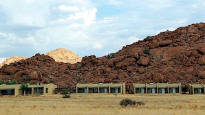Una hilera de edificios modernos de una sola planta con techos planos, ubicados en un paisaje árido y abierto, con una gran montaña escarpada al fondo.