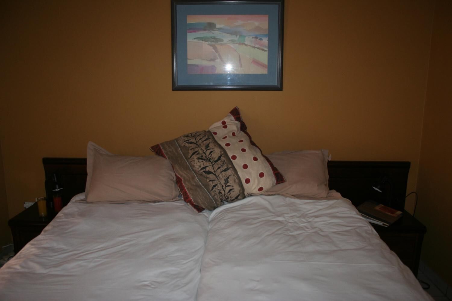 A bed with white bedding and multiple pillows, a framed picture on the wall, and a bedside table with a book and a bottle.