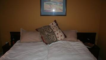 A bed with white bedding and multiple pillows, a framed picture on the wall, and a bedside table with a book and a bottle.