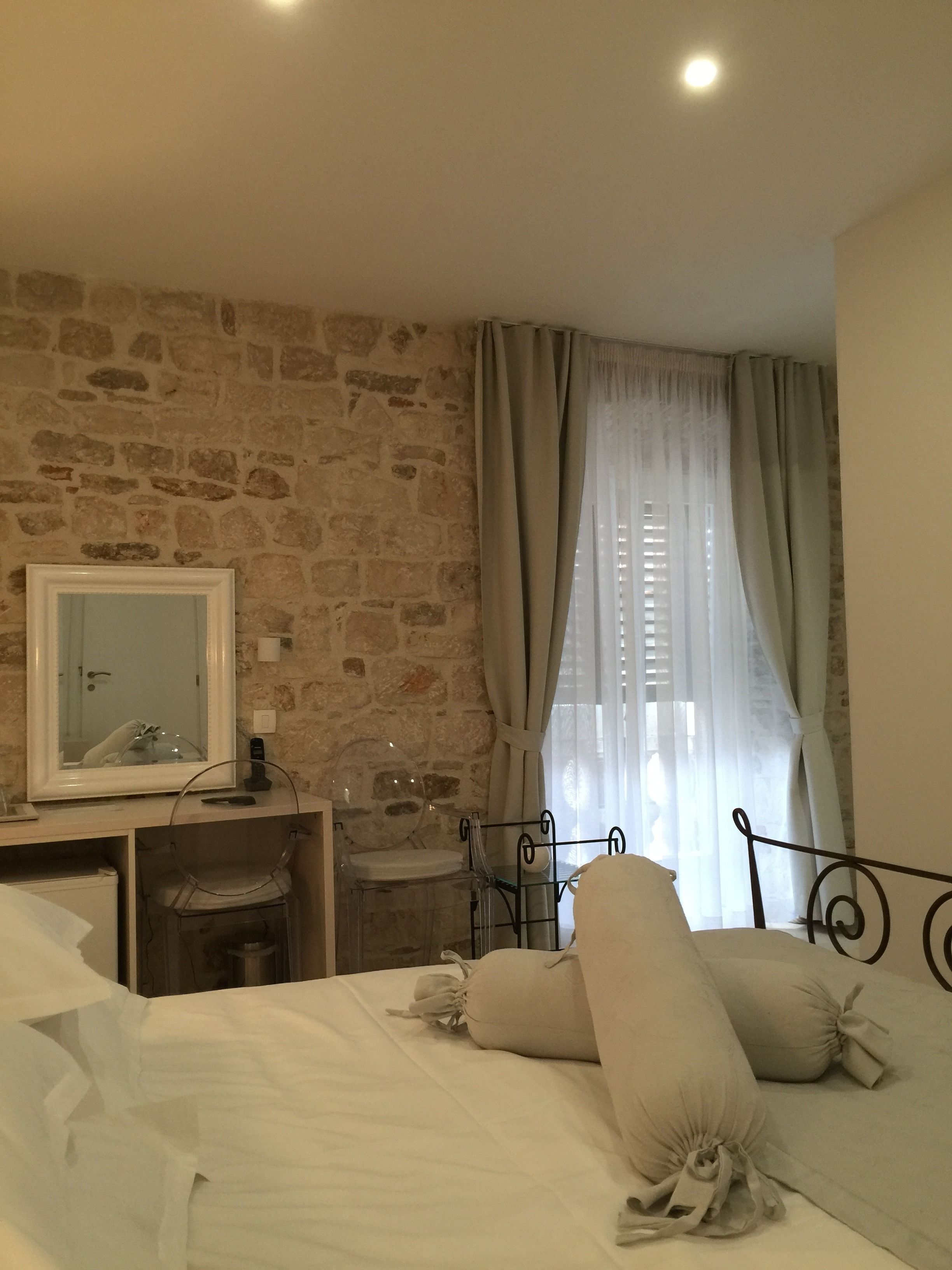A bedroom with a stone wall, a bed with white linens and pillows, a bedside table with a mirror, and a window with sheer curtains.