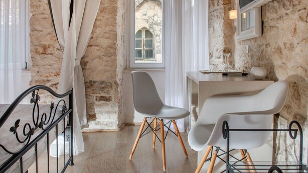A room with a stone wall, a window with a view, a white desk, a white chair, and a bed with a metal frame. - Palace Tartaglia (Split)