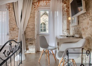 Une pièce avec un mur en pierre, une fenêtre donnant sur un beau paysage, un bureau blanc, une chaise blanche et un lit avec un cadre en métal.