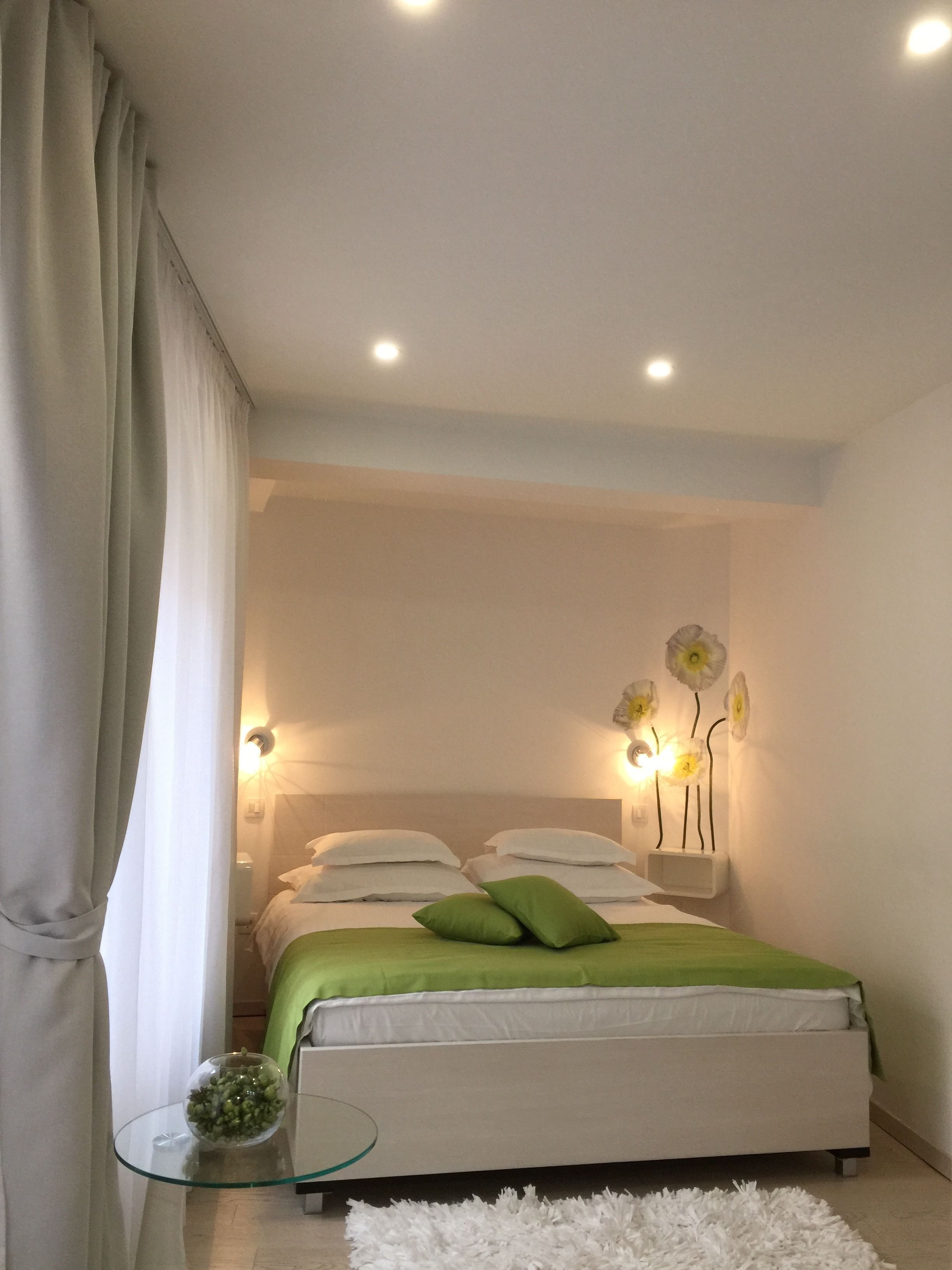 A modern bedroom with a bed, green pillows, a glass table with a flower vase, and wall-mounted lights.