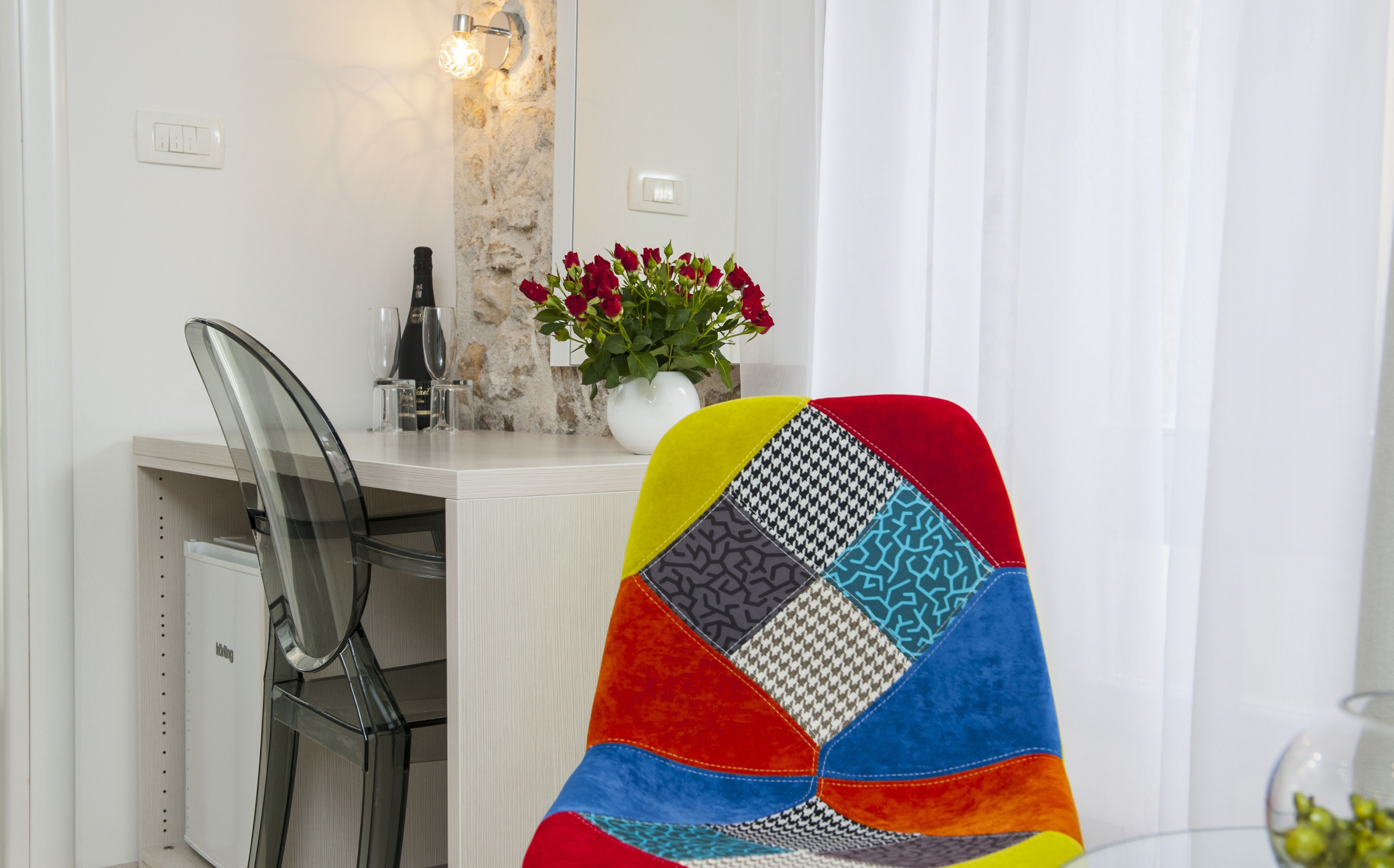 A dining area with a white table, a chair with a colorful quilted backrest, a vase with red flowers, and a wall-mounted light fixture.