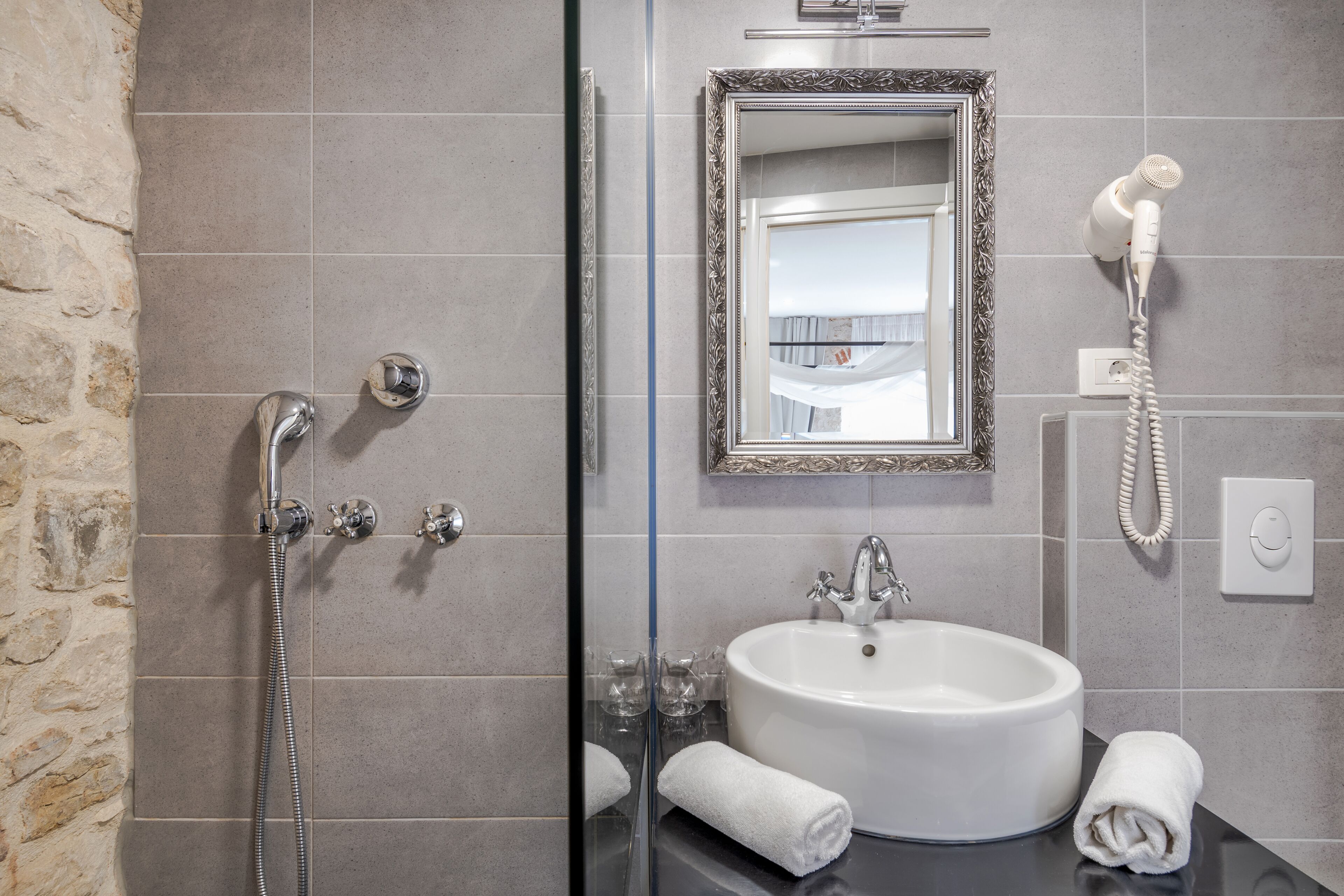 A bathroom with a glass-enclosed shower, a white sink, a mirror, and a hairdryer on the wall.